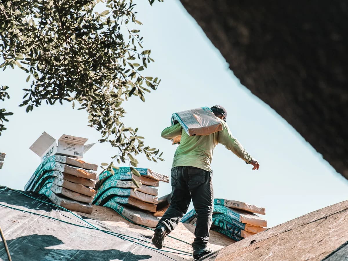 Roofer carrying a bundle of shingles across a roof past stacked bundles