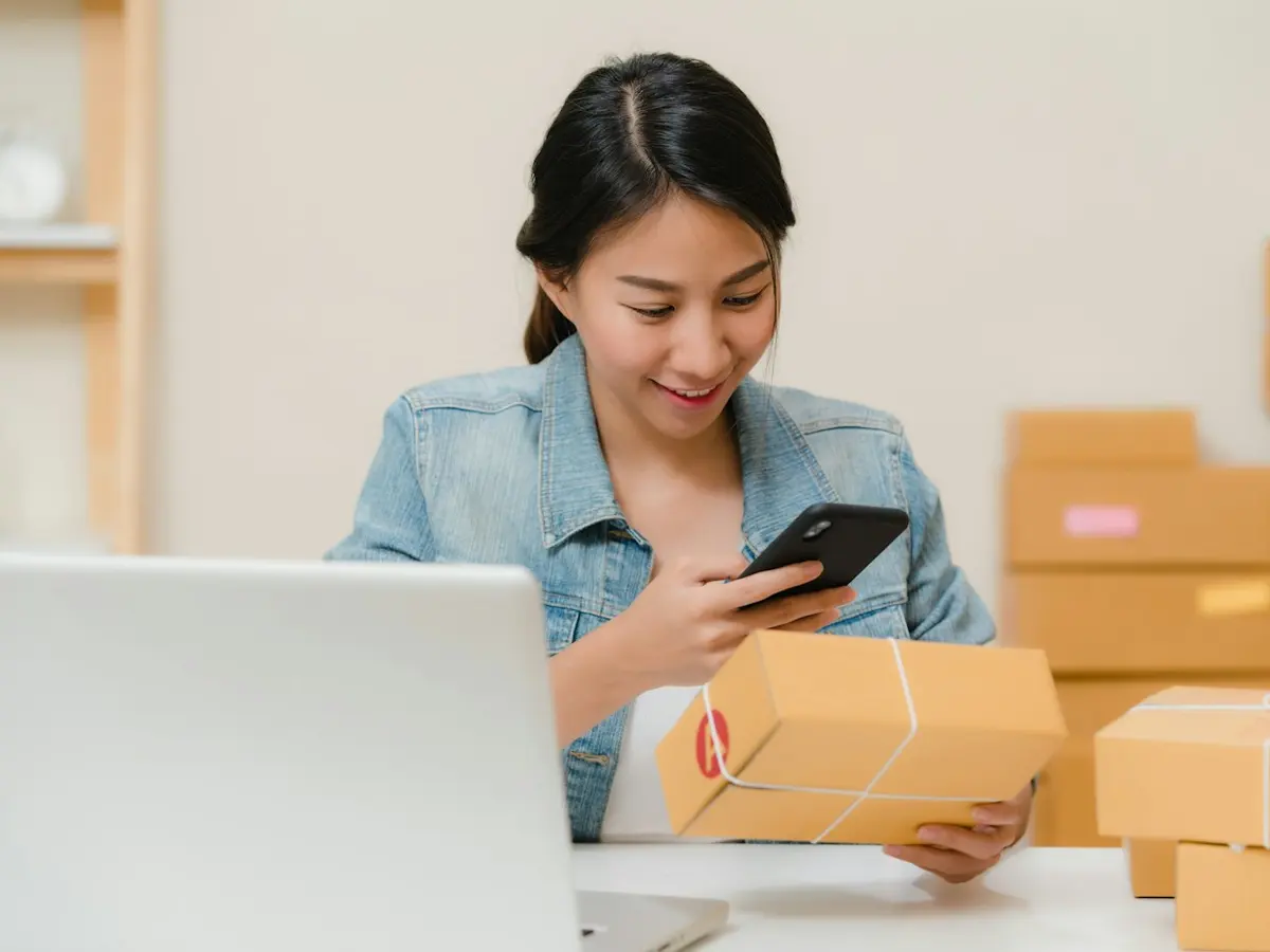 Online seller checking an order on her phone beside packed shipping boxes