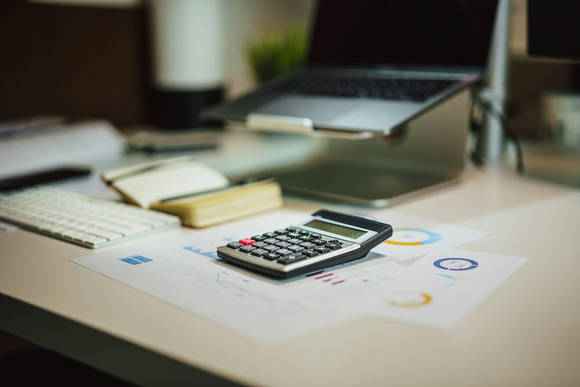 A calculator resting on printed financial charts beside a laptop